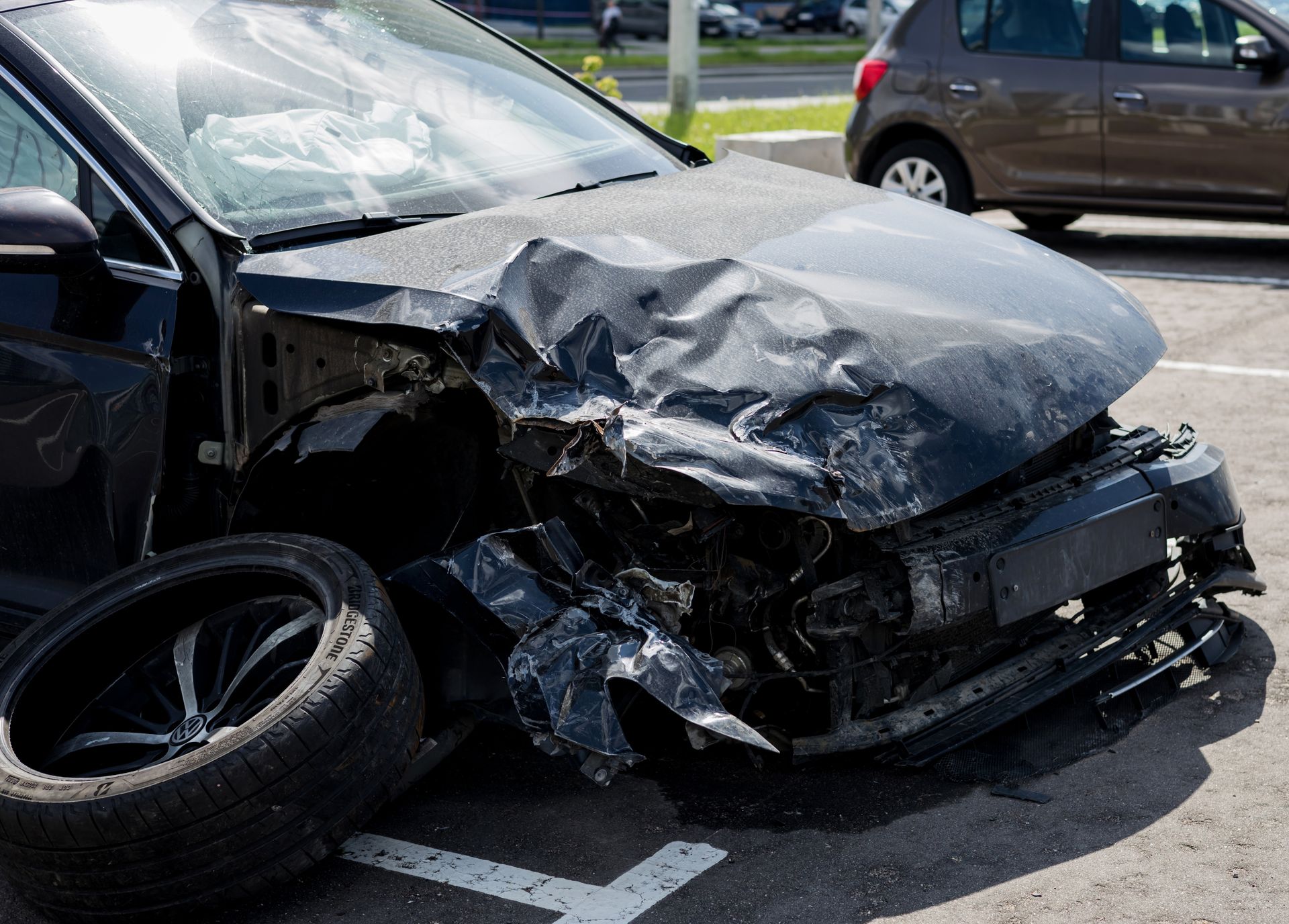 Damaged black car with severe front-end collision in a parking lot, tire detached.