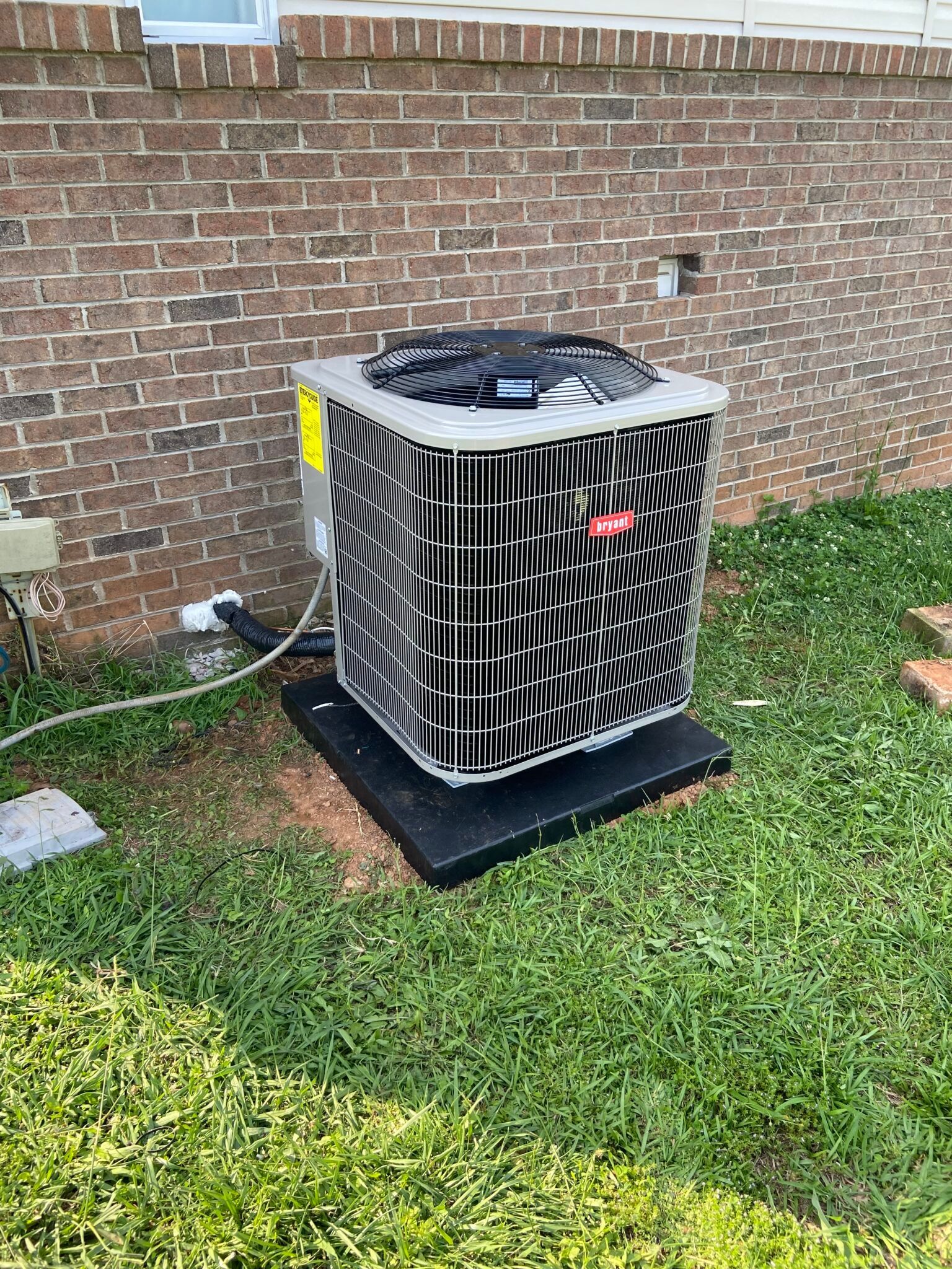 An air conditioner is sitting in the grass next to a brick wall.