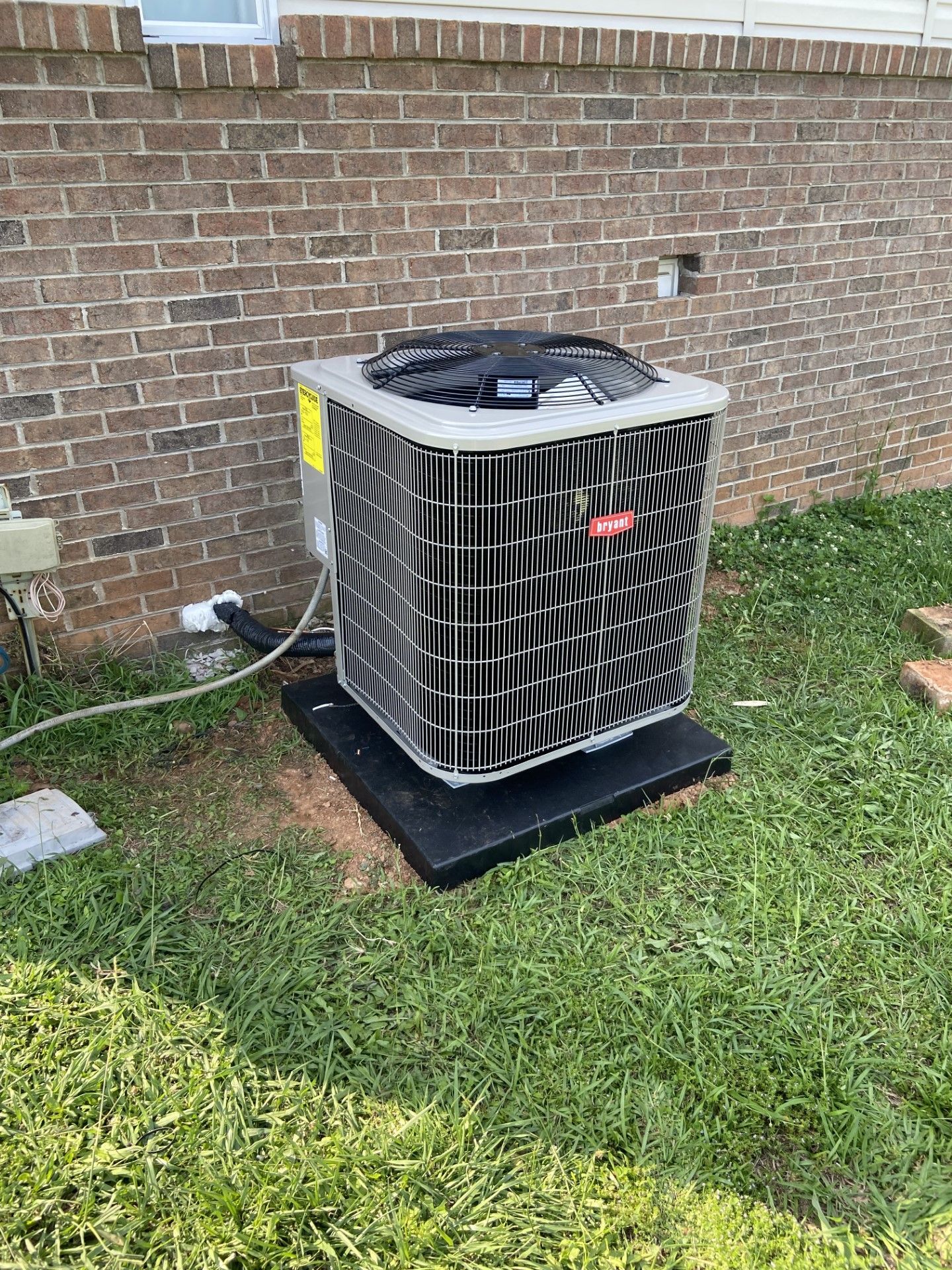 An air conditioner is sitting in the grass next to a brick wall.