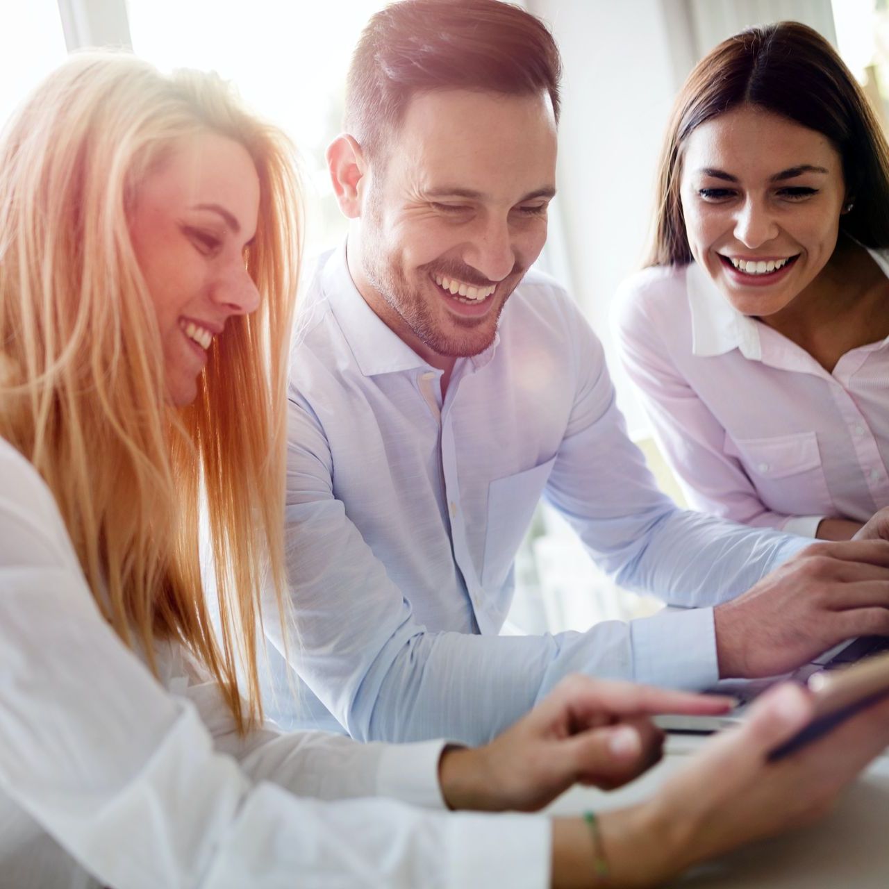 Three people smiling while looking at a mobile phone and laptop together in an office.