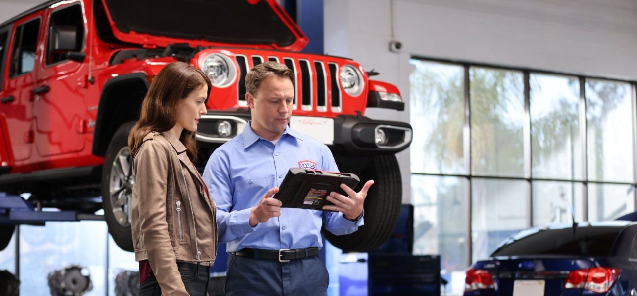 Mechanic showing a woman a tablet next to a red Jeep in a car repair shop.