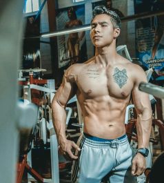 Fit person posing shirtless in a gym; wolf tattoo, six-pack abs, athletic build.