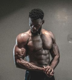 Muscular person flexing arms, chest, and abs; indoors, neutral background, looking down.