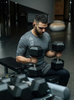 Man in gray shirt seated, lifting black dumbbells on a gym bench.