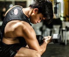 Man in gym curls dumbbell. He wears a black vest and shorts.