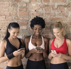 Three women in athletic wear using smartphones near a brick wall, smiling.