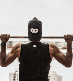 Person doing a pull-up, wearing a black hat and tank top with an infinity symbol. Gray background, showing arms.