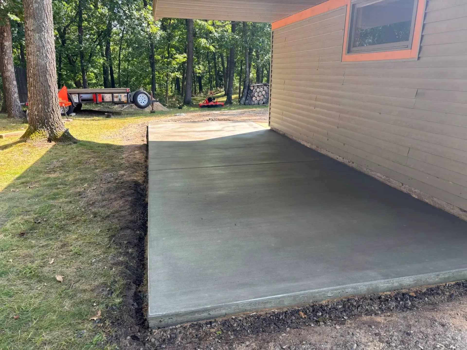 A newly poured concrete patio slab adjacent to a house exterior, with trees and a trailer visible in the background.