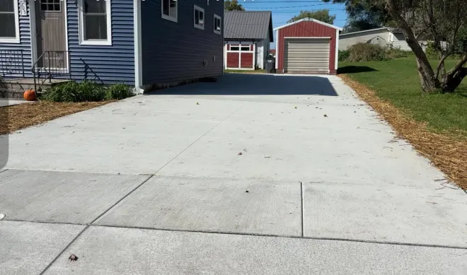 A concrete driveway leads toward a house with blue siding and a detached red garage under a clear sky.
