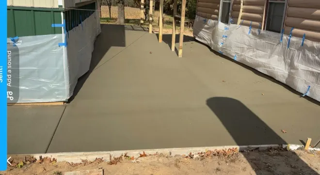 Newly poured gray concrete patio between a green structure and a log cabin house under construction.
