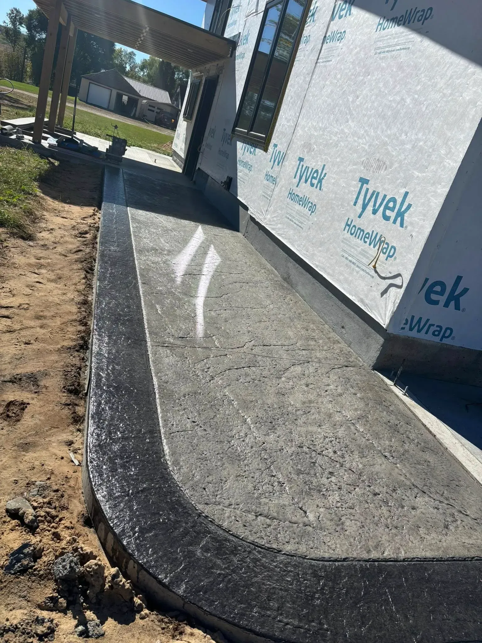 A freshly poured concrete sidewalk with a smooth black border runs along the side of a building wrapped in Tyvek.