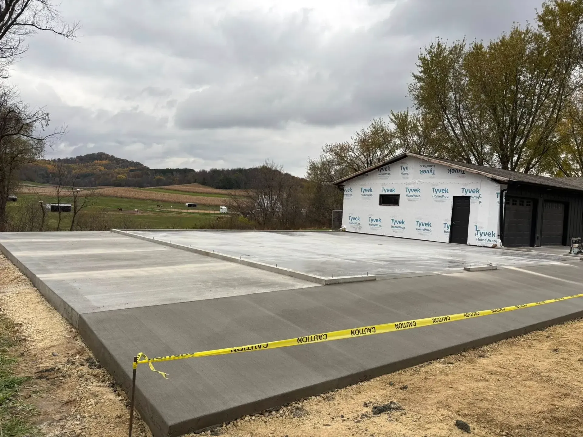 A newly poured concrete foundation slab sits in front of a white building under construction on a cloudy day.