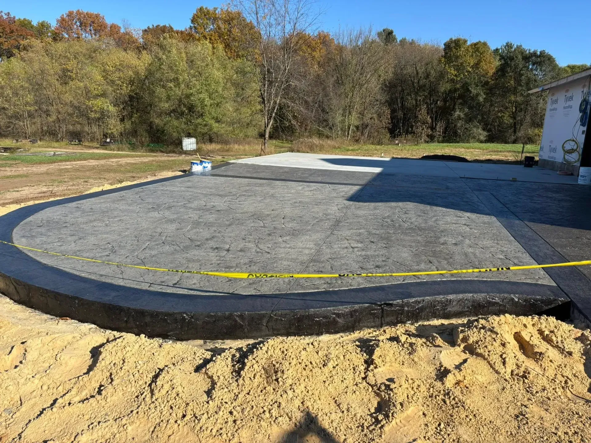 A newly poured concrete patio with a dark border and stamped texture, surrounded by dirt and an autumn woodland backdrop.