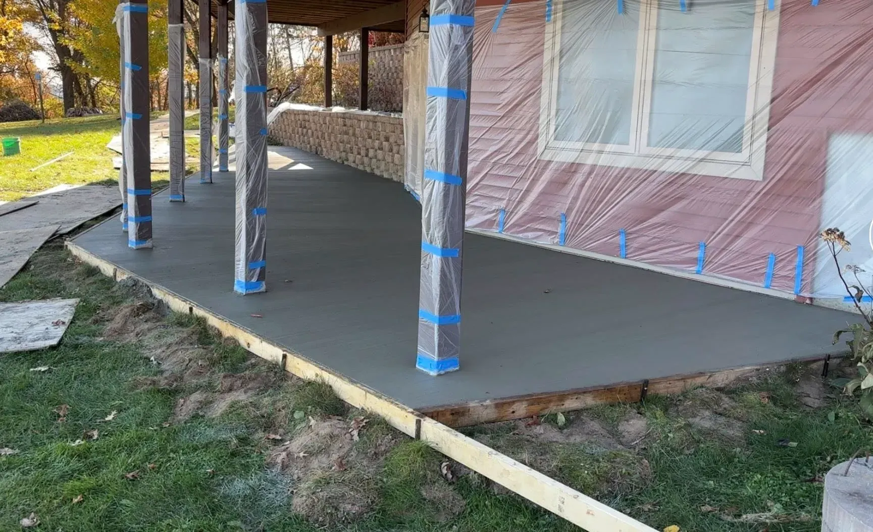 A newly poured concrete patio wraps around a house with columns wrapped in protective plastic.