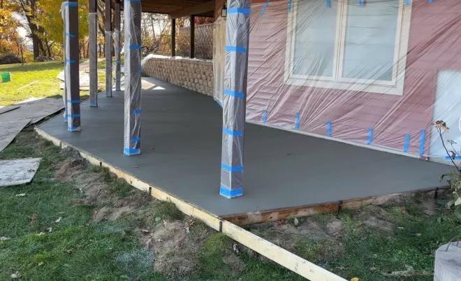 A freshly poured concrete porch under construction with wrapped support pillars and a house wall covered in plastic.