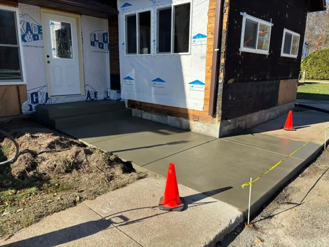 Freshly poured concrete sidewalk and steps leading to a house exterior undergoing renovation, marked by orange cones.