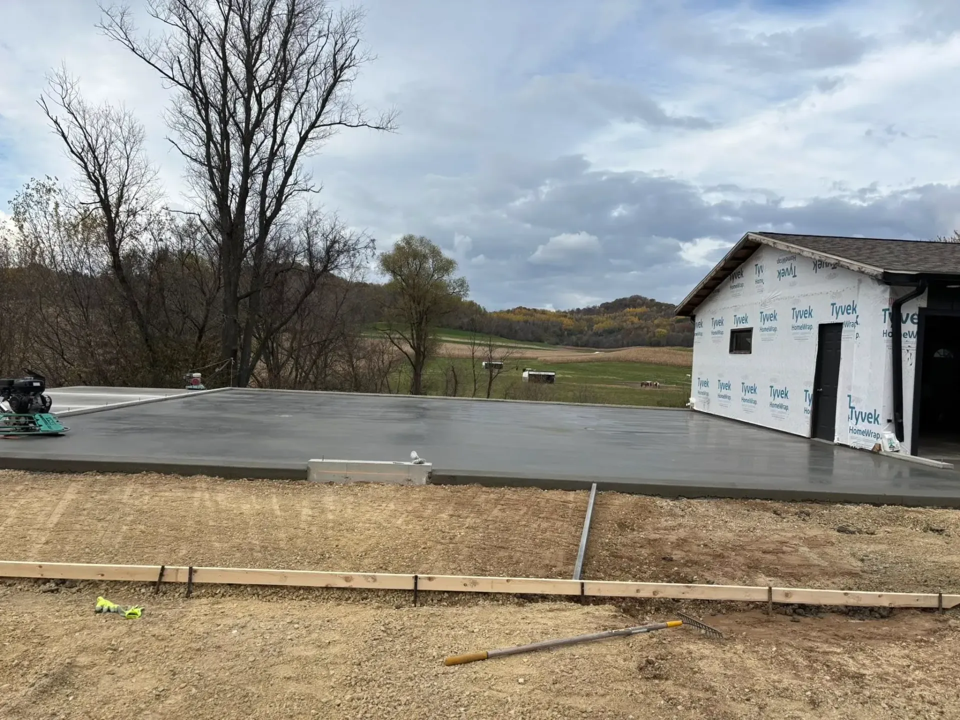 A freshly poured concrete slab extends from an unfinished building under construction in a rural, grassy field.