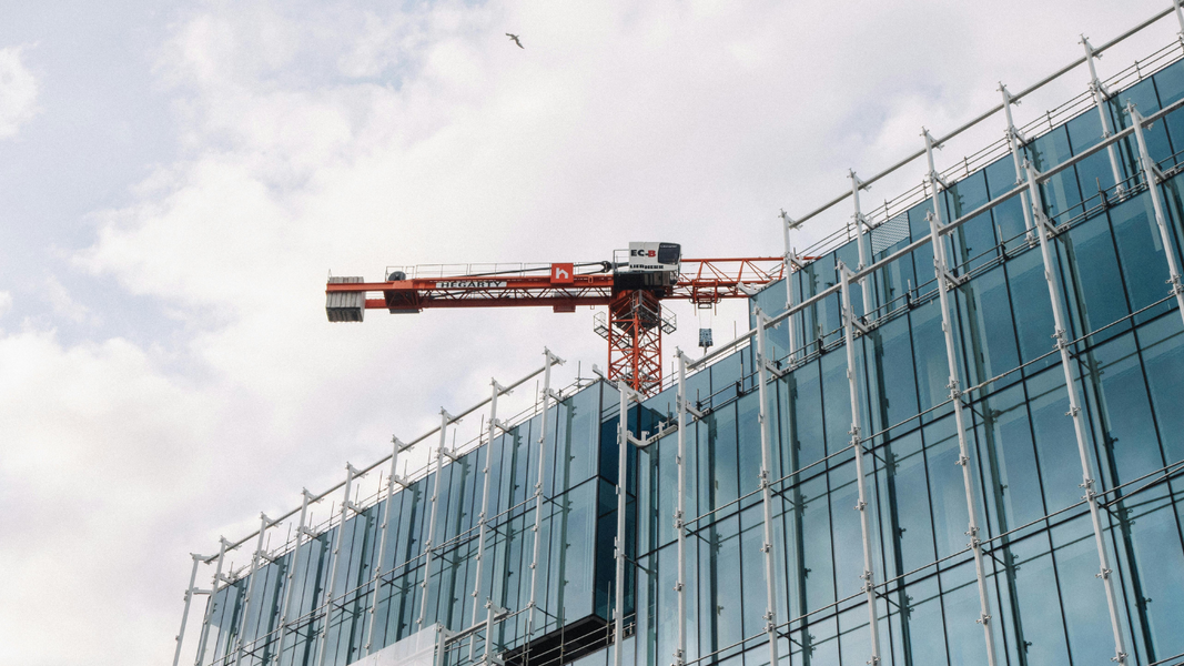 An orange construction crane stands atop a modern building featuring a glass facade and surrounding scaffolding.