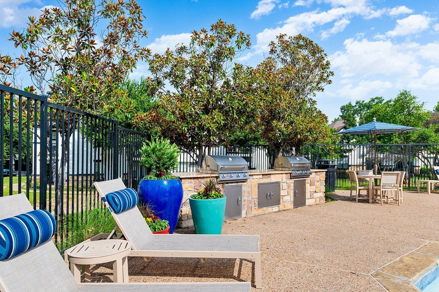 Poolside grill area with stainless steel grills, lounge chairs, tables, and shade umbrellas under a blue sky at Villas of Vista Ridge in Lewisville, TX.