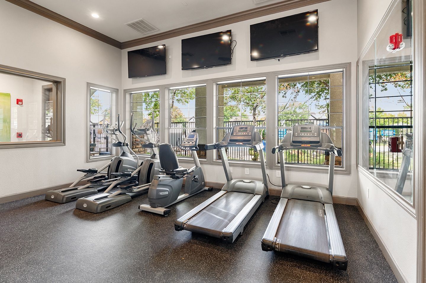 Gym with cardio machines near windows, TVs on the wall, and a mirrored wall section at Villas of Vista Ridge in Lewisville, TX.