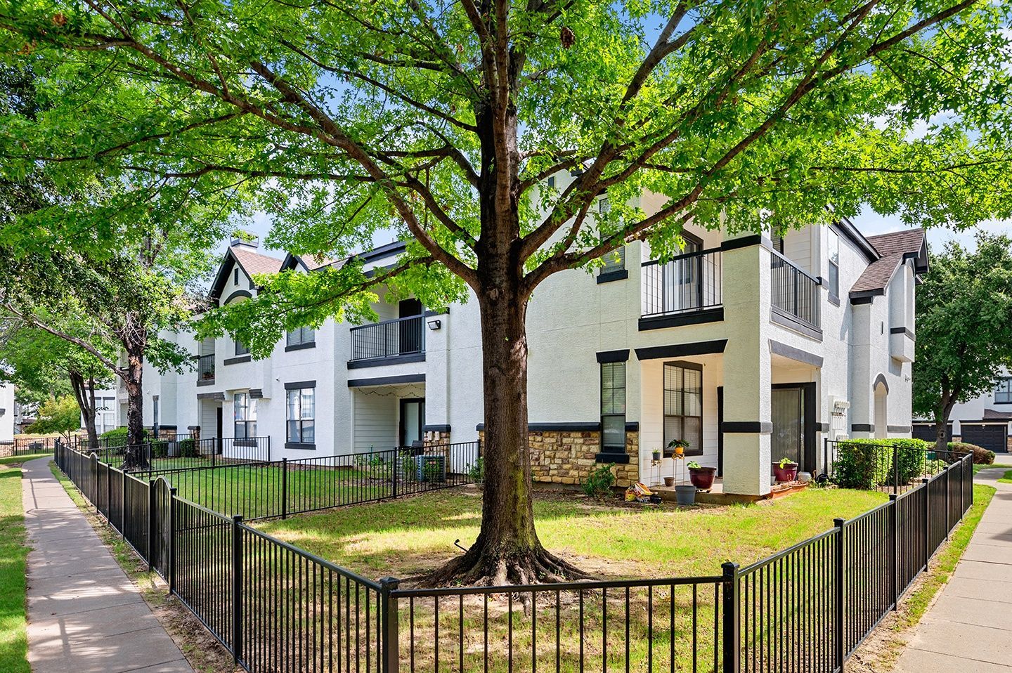 Apartment building with white stucco walls, black accents, and wrought iron fence, shaded by a large tree at Villas of Vista Ridge in Lewisville, TX.