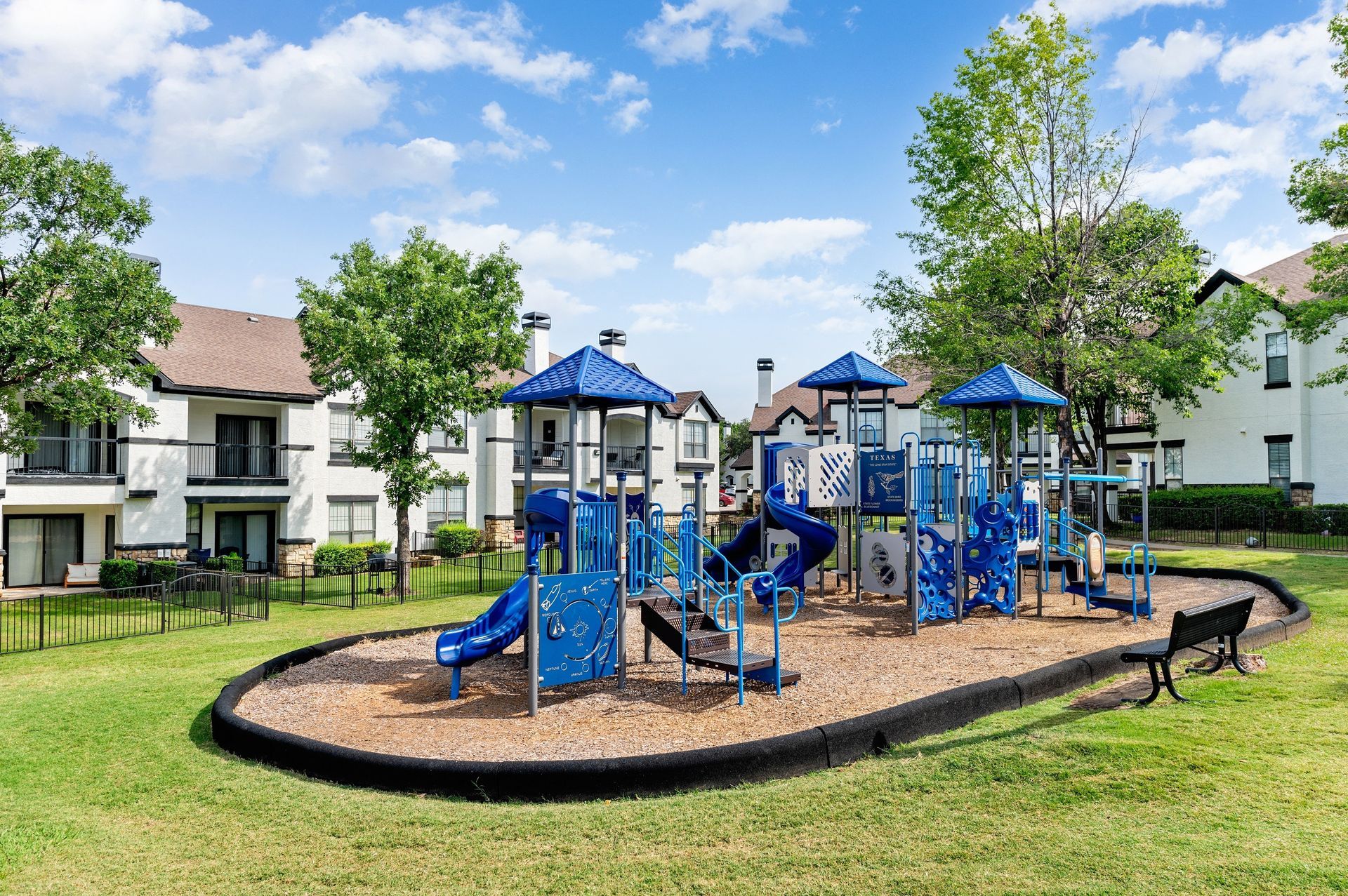 Playground area surrounded by greenery and apartment buildings at Villas of Vista Ridge in Lewisville, TX.