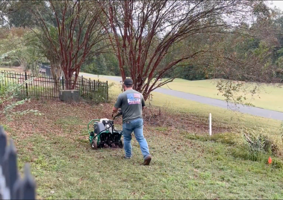 Man using a tiller on a hillside lawn near trees and a golf course.