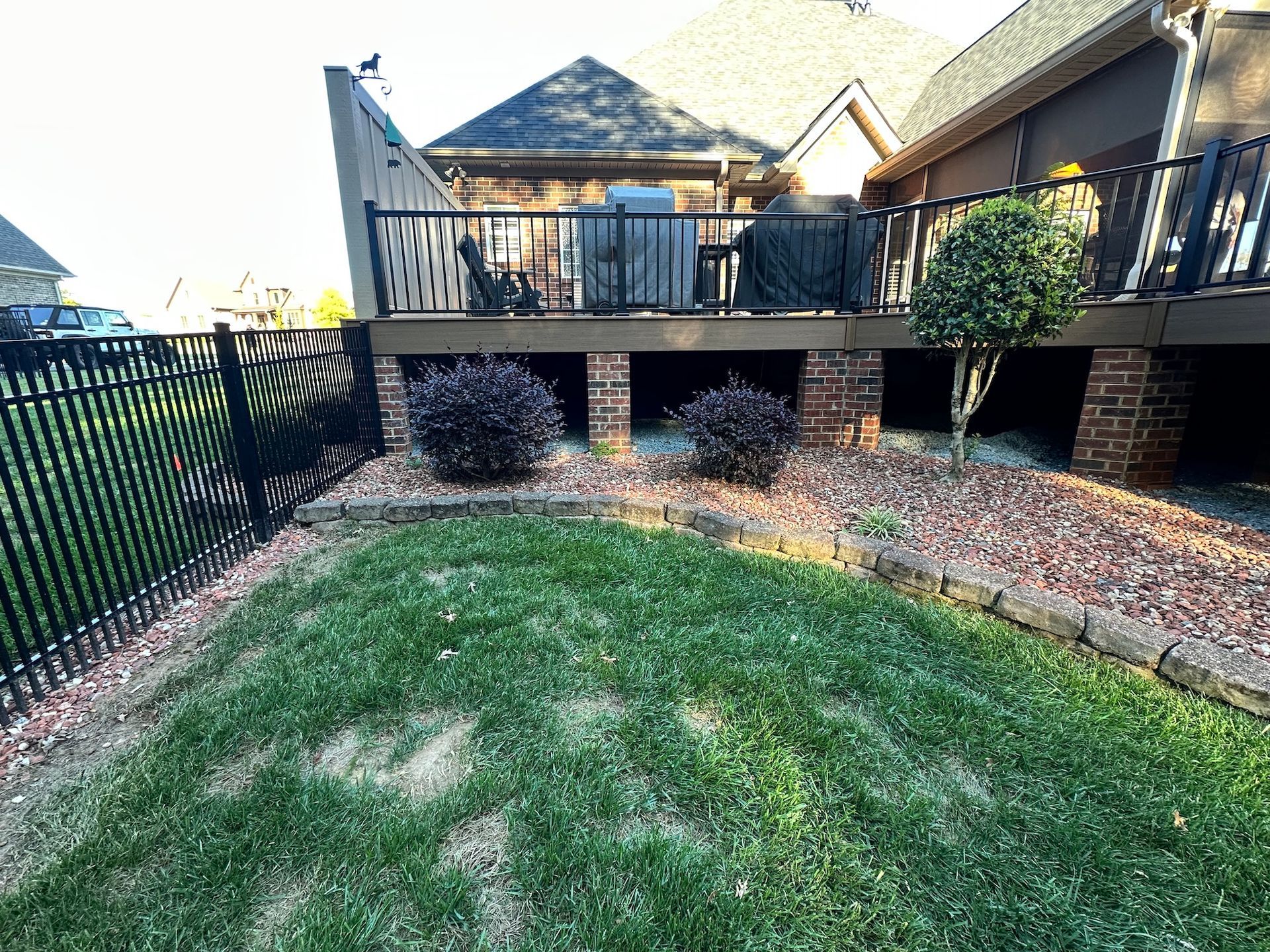 Backyard deck with black railing and fence, red brick supports, shrubs, and a grassy lawn.