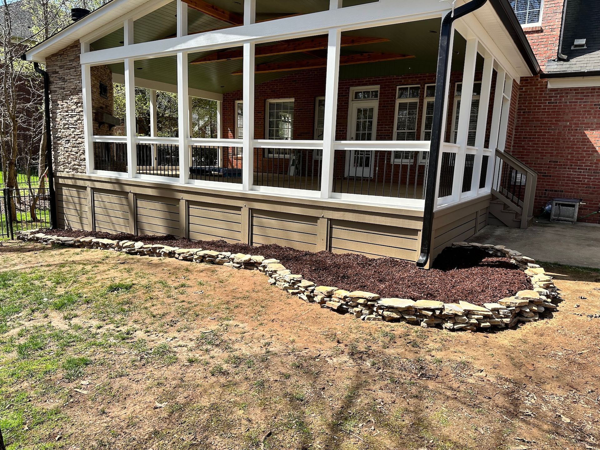 Screened porch with brown trim, surrounded by a rock border filled with mulch, next to a brick house.