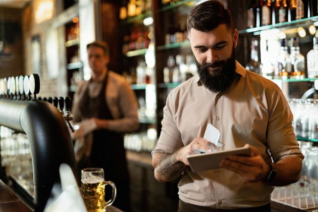 A man with a beard is using a tablet in a bar.