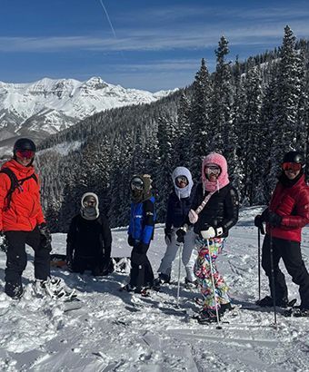 Group of skiers on snowy mountain, wearing colorful ski outfits and face coverings, with mountains and trees in background.