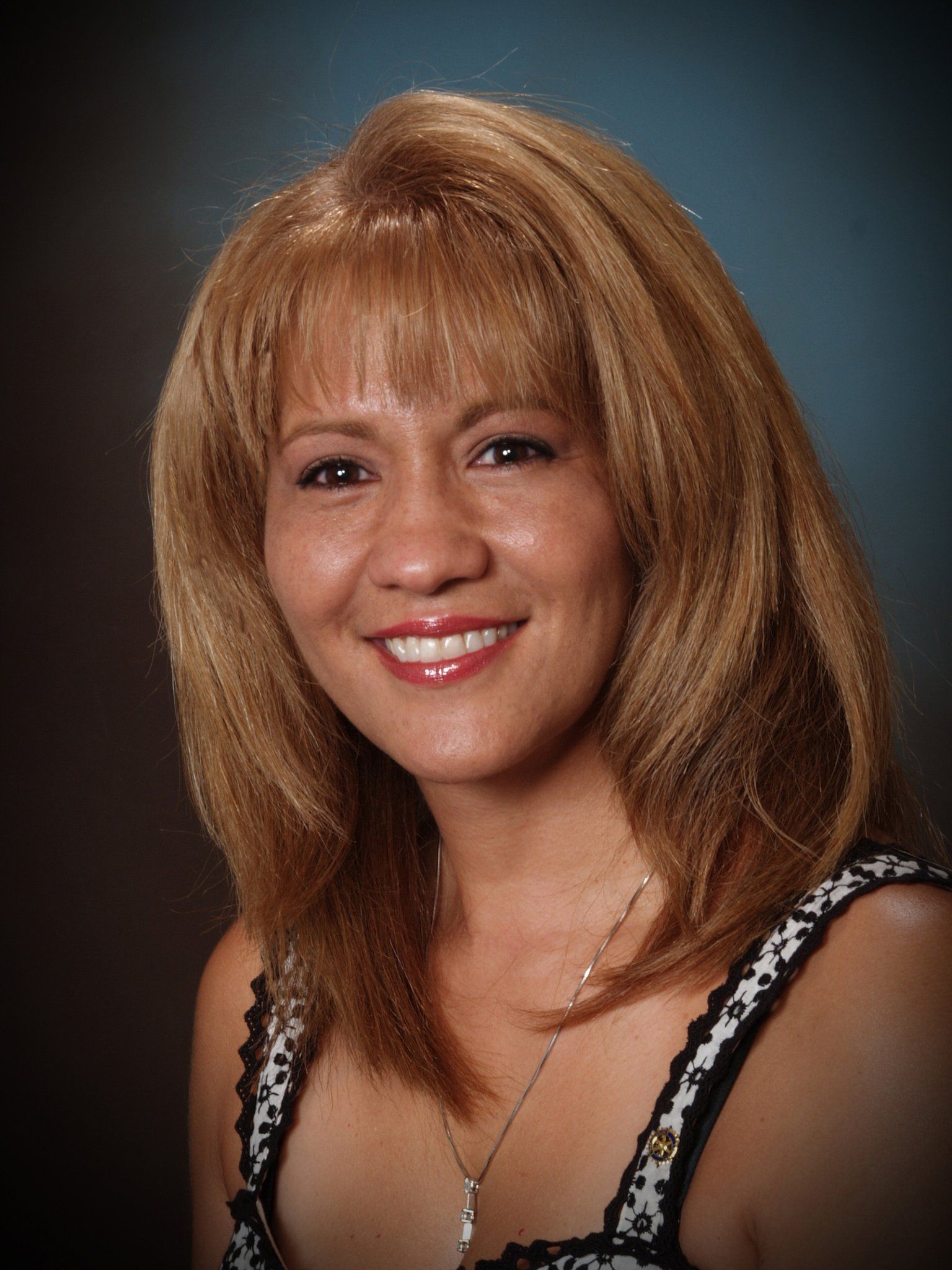 Smiling woman with shoulder-length blonde hair and bangs, wearing a patterned tank top. She has a necklace and is against a dark blue backdrop.