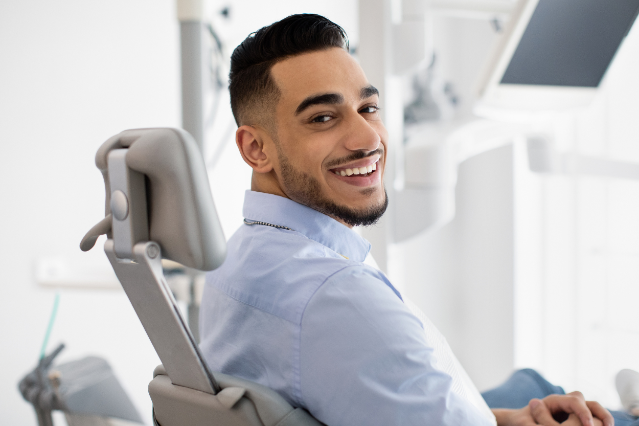 A smiling man with dark hair and a beard sits in a dentist's chair. He looks back toward the viewer.