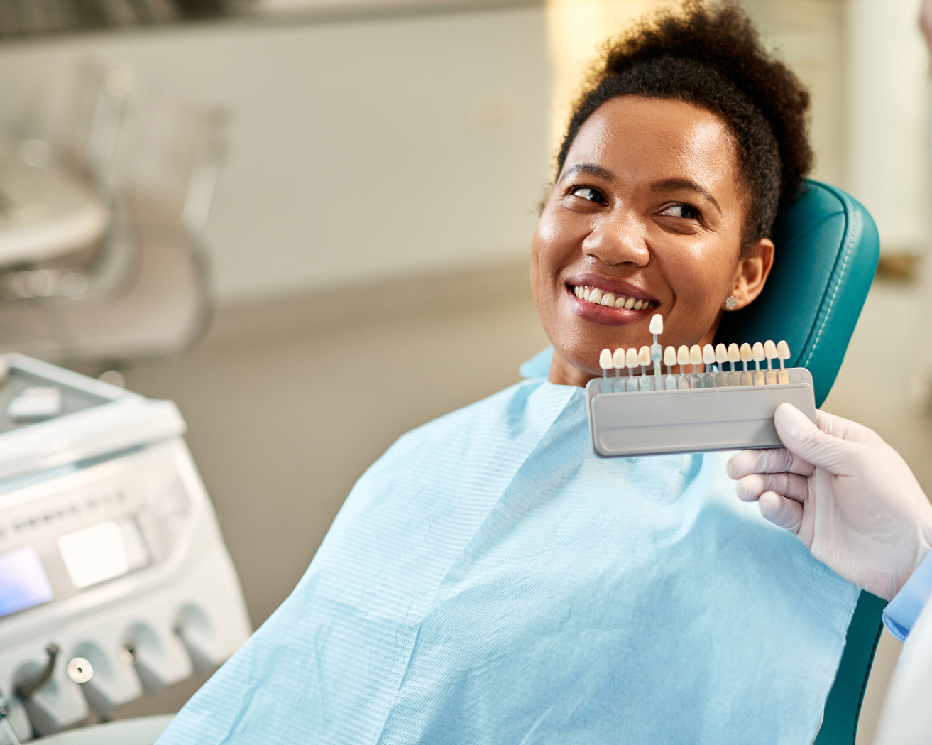 Woman at dentist smiles while shade samples are compared to her teeth. Clinic setting.