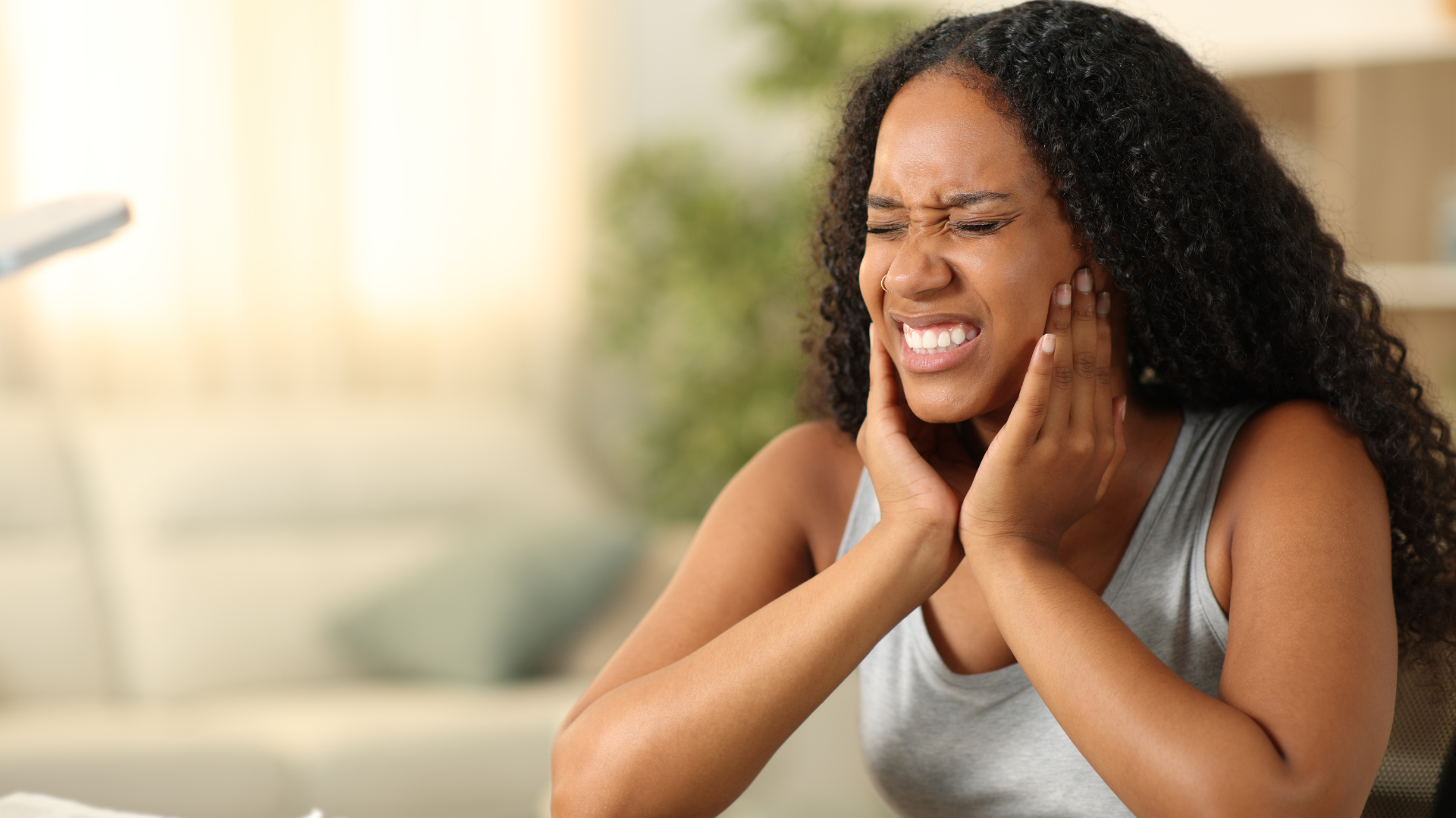 Woman with dark curly hair, clutching her face in pain, indoors, with pained expression.