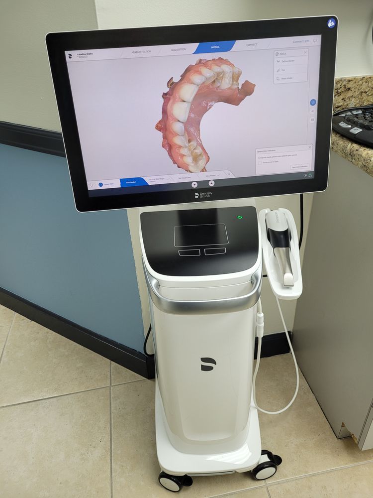 A white dental scanner displaying a 3D model of teeth on a screen. The device is on wheels, in a dentist's office setting.