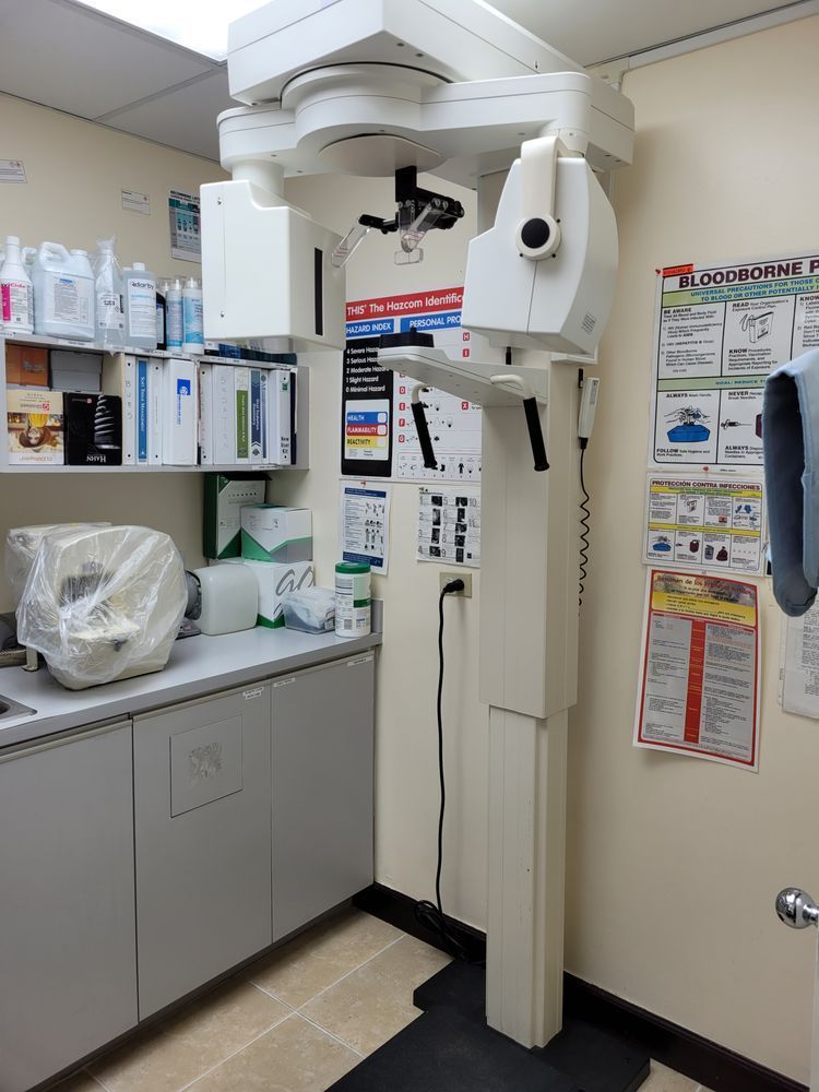 Dental panoramic X-ray machine in a clinical setting. It's tall, white, and stands on a black base next to a counter with medical supplies.