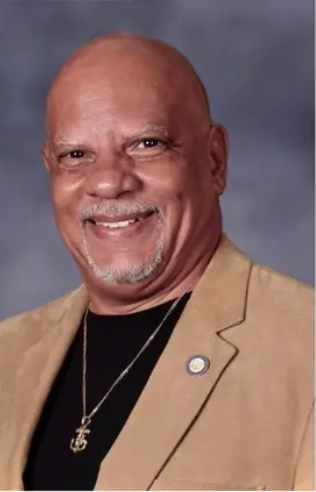 A bald man wearing a tan jacket and a black shirt