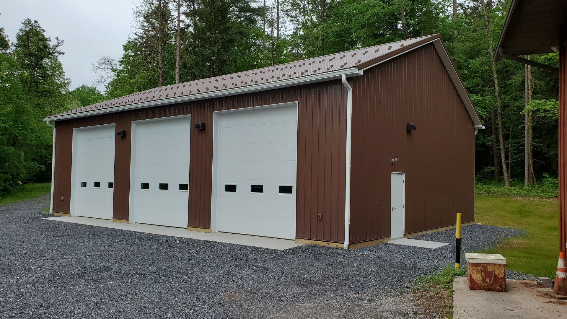 A brown garage with white doors is sitting in the middle of a gravel driveway.