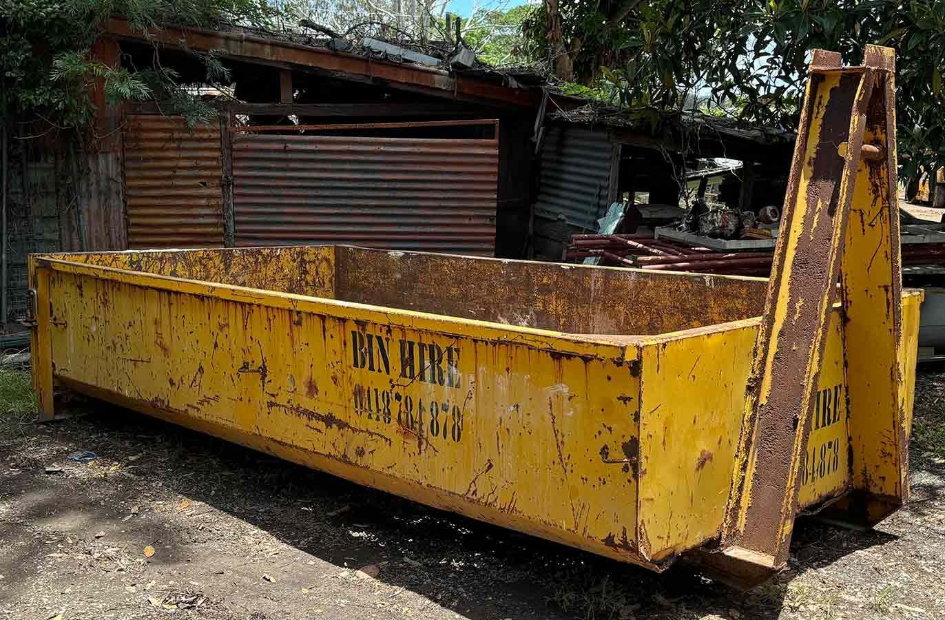 A large yellow dumpster is sitting in the dirt in front of a building.