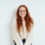 A woman with red hair and glasses is wearing a white sweater and smiling.