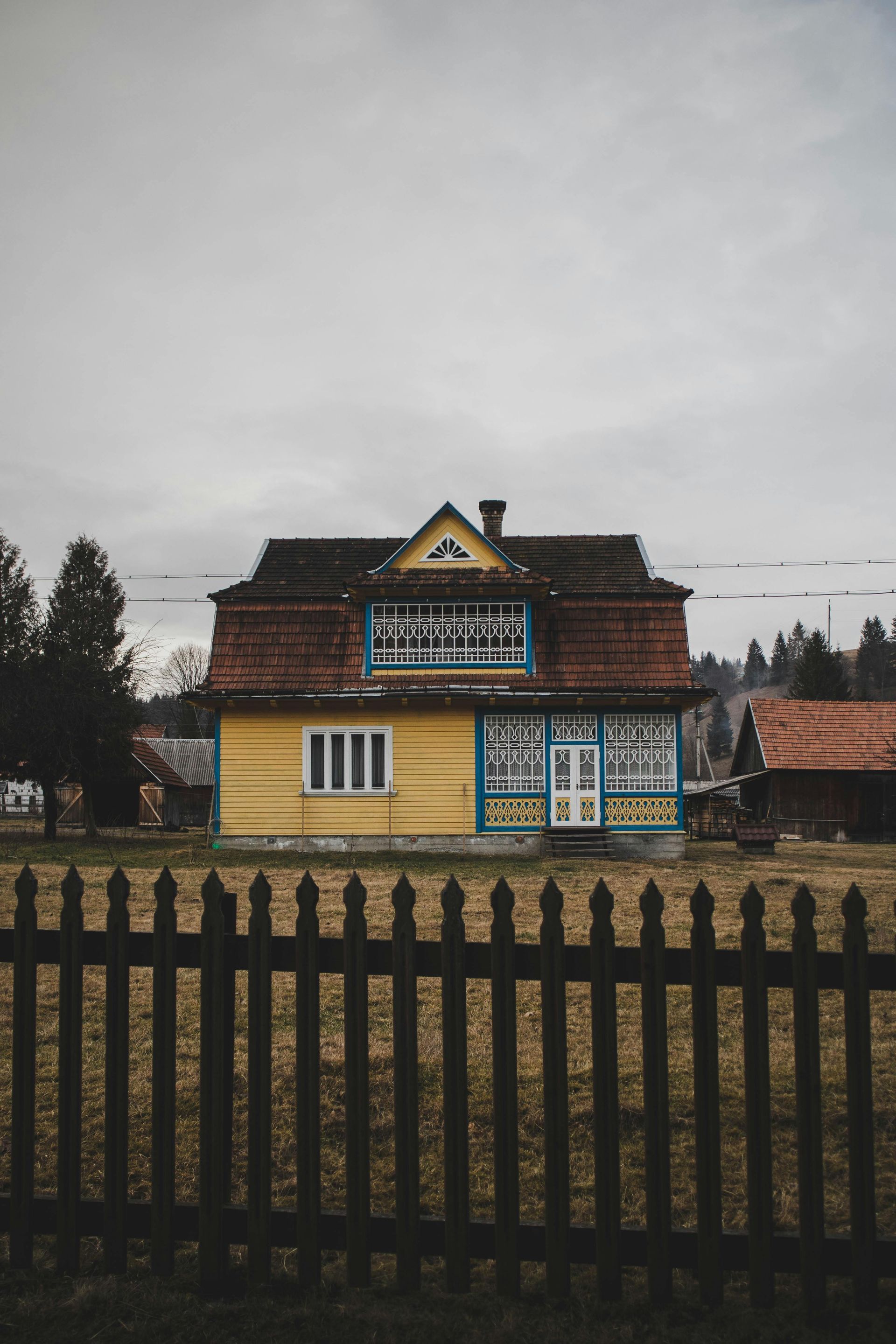 A yellow house with a tiled roof behind a black picket fence under an overcast sky.