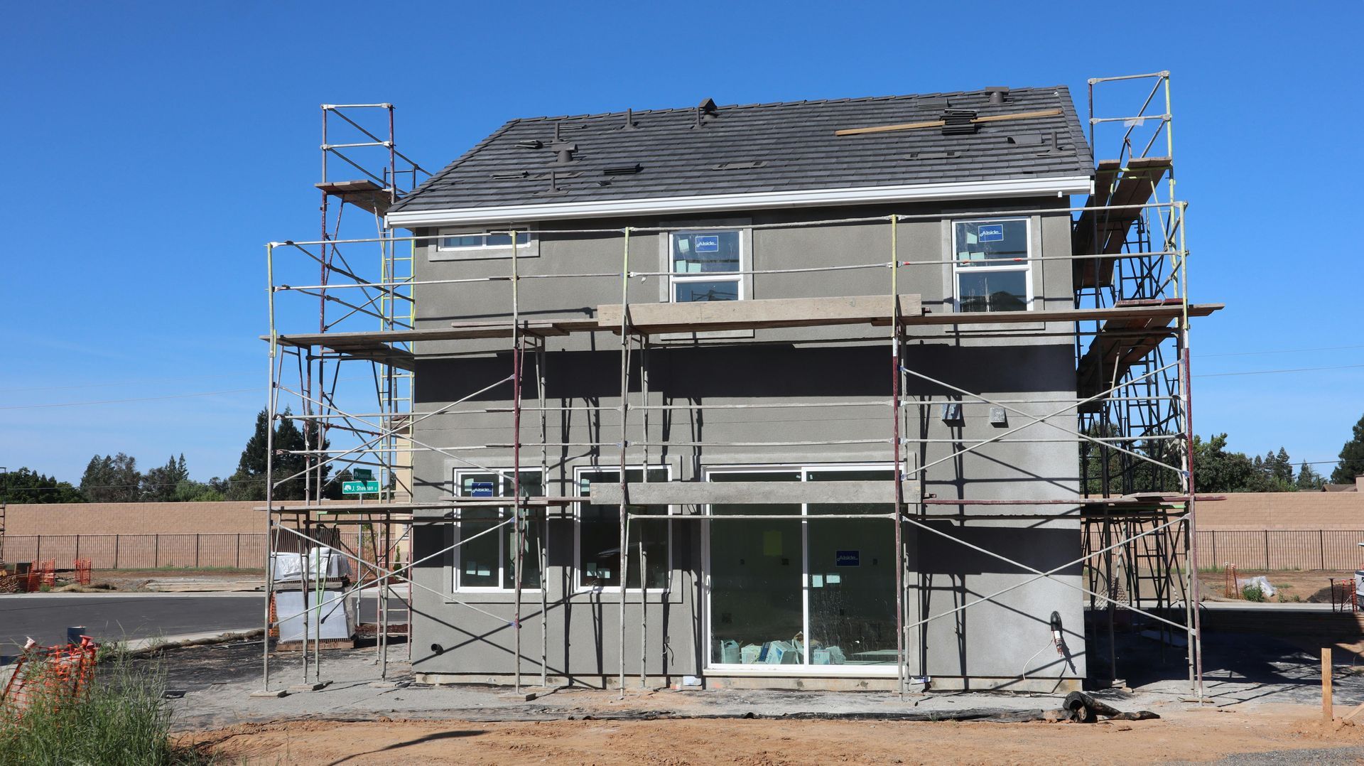 Two-story house under construction with scaffolding, gray siding, and unfinished exterior in a sunny lot