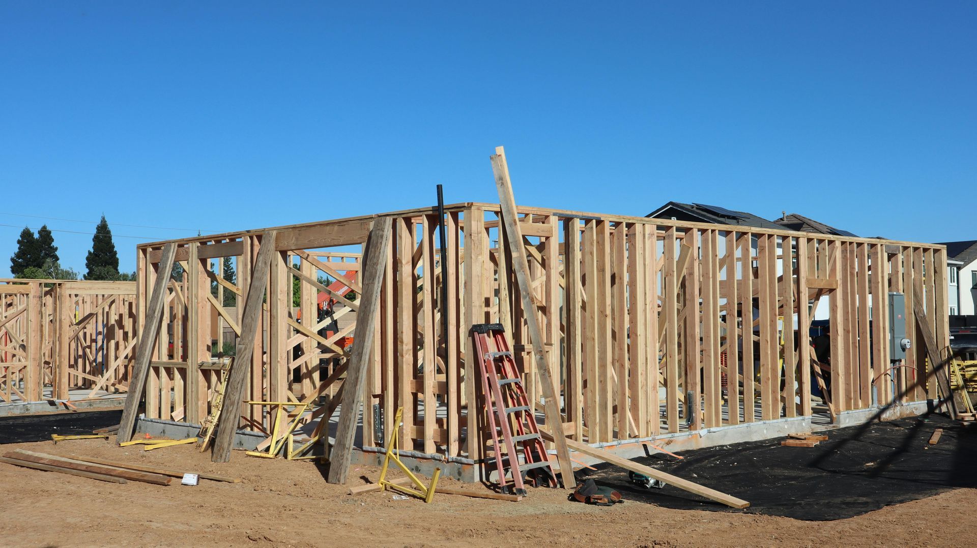 Wood-framed house under construction on a dirt lot, with ladders and scattered building materials.