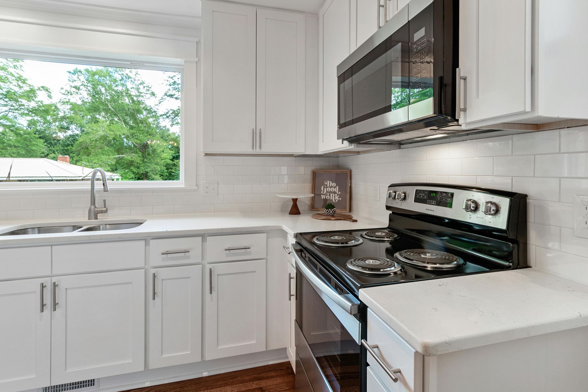 Bright white kitchen with stainless steel stove, microwave, sink, and large window above the counter