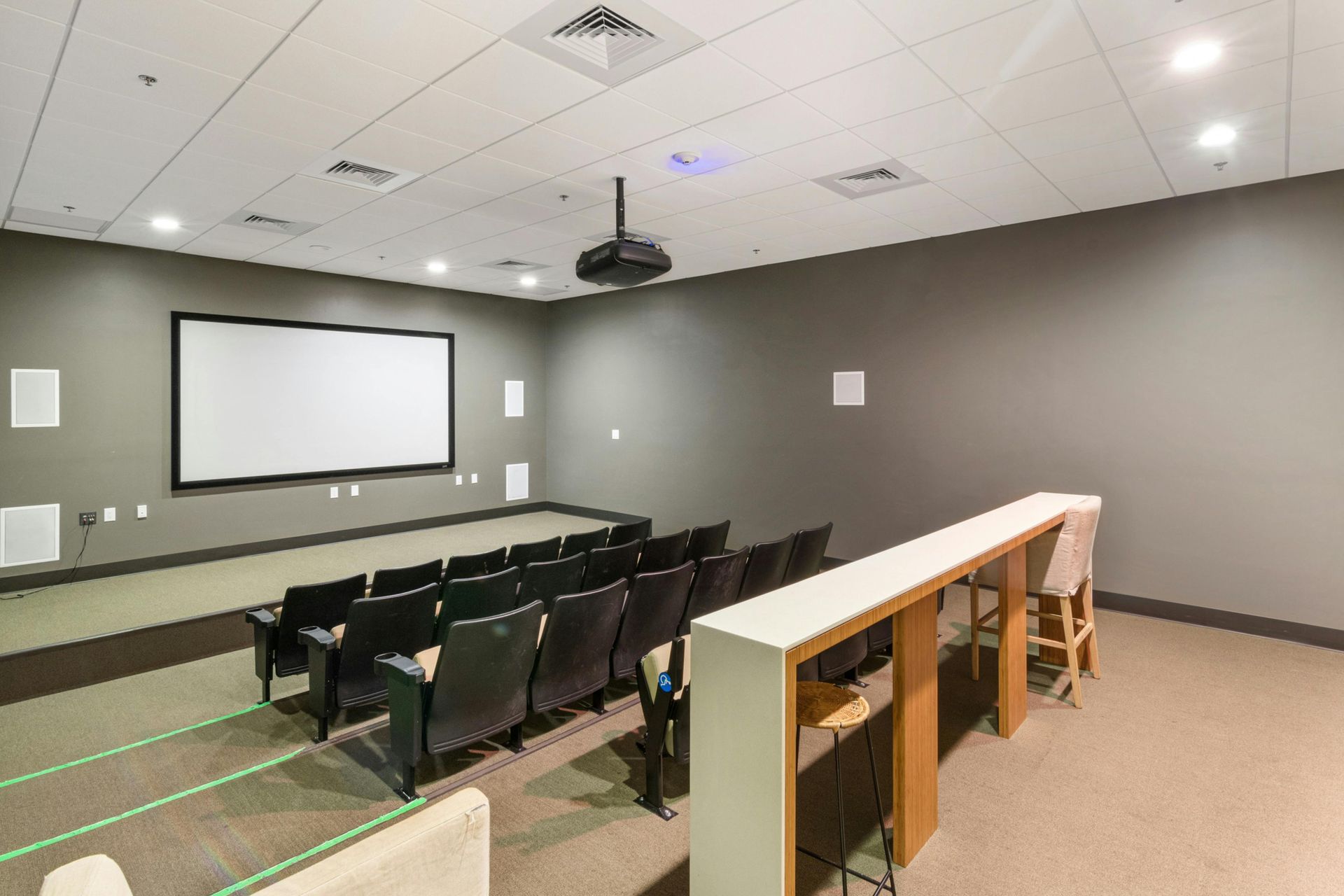 Empty lecture room with projector screen, rows of black chairs, and beige walls and carpet.