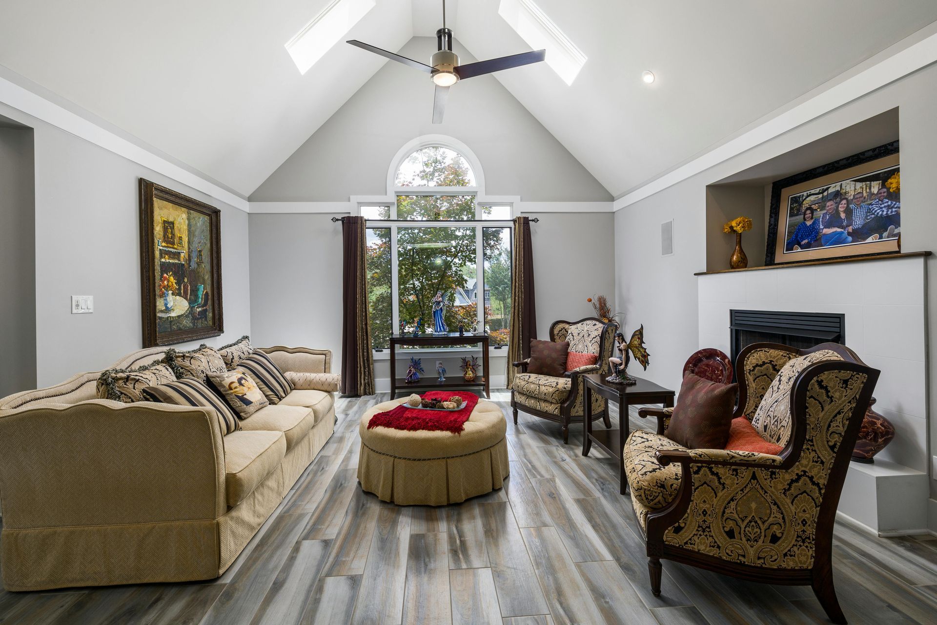 Bright living room with vaulted ceiling, patterned chairs, sofa, and a round ottoman facing tall windows.