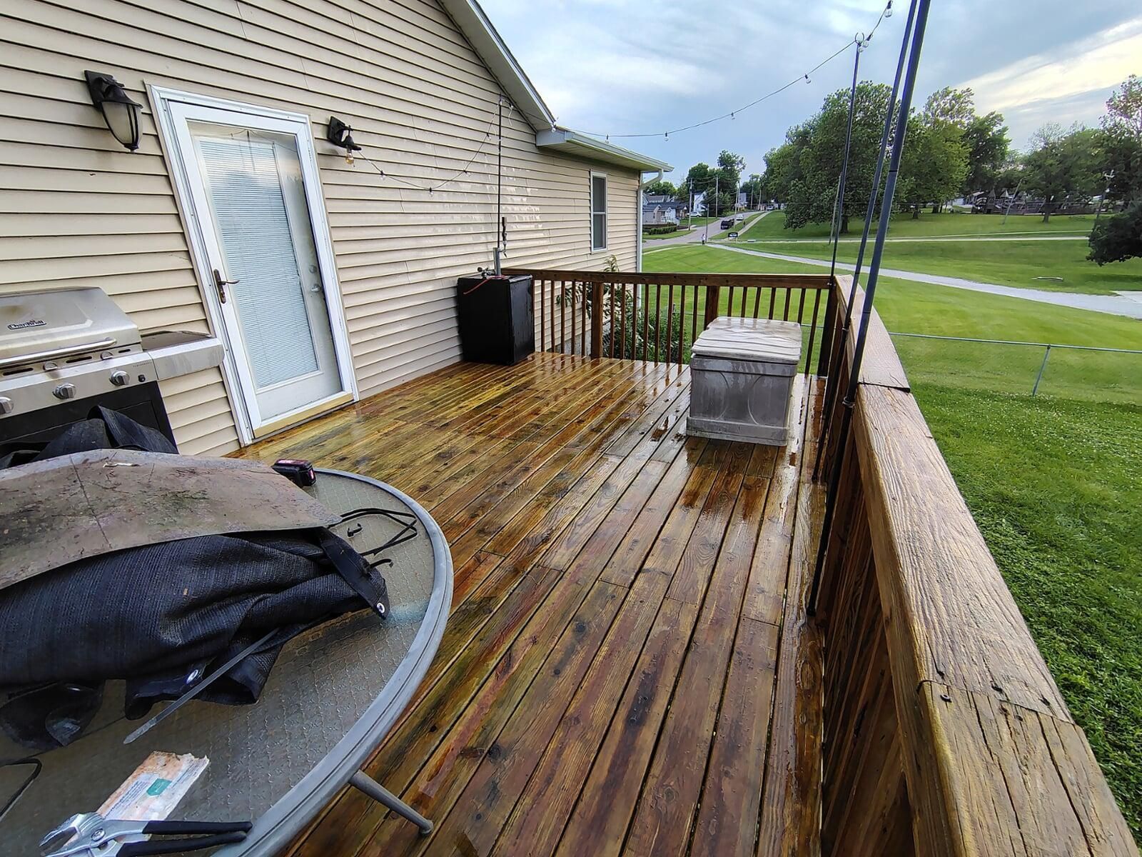 A wooden deck with wet, stained planks extends from a beige house, alongside a grassy yard. A grill, furniture, and a storage chest are visible.