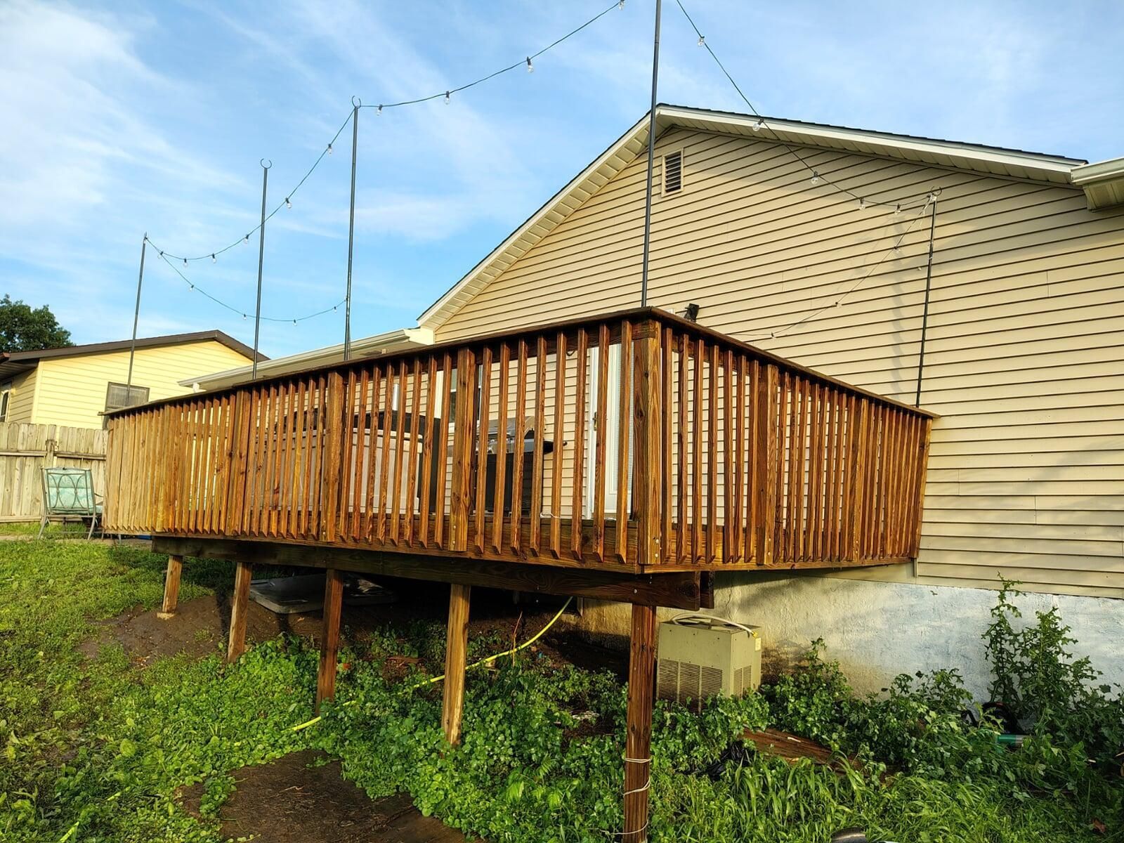 Wooden deck attached to a house with string lights overhead. The deck is elevated with vertical supports.