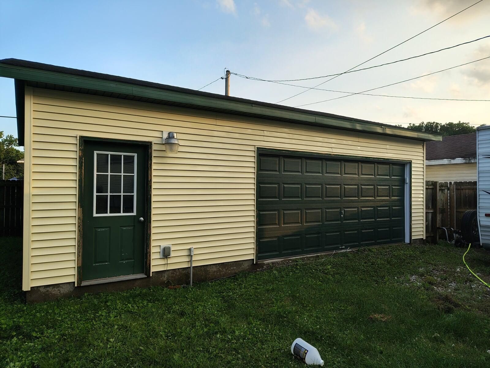 Yellow-sided detached garage with a green door and garage door. Dark green roof and trim.